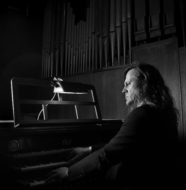 A music director sitting at a piano, composing a melody, with a focused expression and a notepad beside them. The room is dimly lit, creating a contemplative atmosphere.