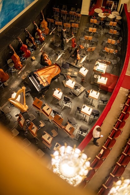 A panoramic view of a live concert featuring an orchestra performing a popular international drama OST. The stage is brightly lit, and the audience is captivated by the performance. The atmosphere is electric and emotional.